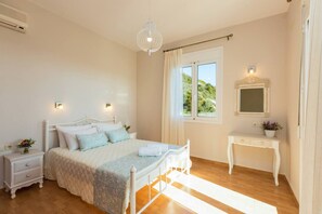 A bedroom with a bed, bedside tables, a dresser, a hanging light fixture, a window with curtains, and a view of greenery outside.