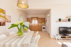 Eine moderne Küche mit Holzschränken, einem weißen Kühlschrank und einem Herd. Es gibt einen Essbereich mit einem weißen Tisch und Stühlen, sowie einen Fernseher auf einem Holzboden.