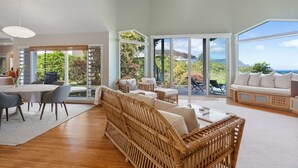 Ein großzügiger Wohnbereich mit einem großen Fenster und Blick ins Freie, ein Essbereich mit Tisch und Stühlen sowie eine Sitzecke mit einem Korbsessel und Kissen.
