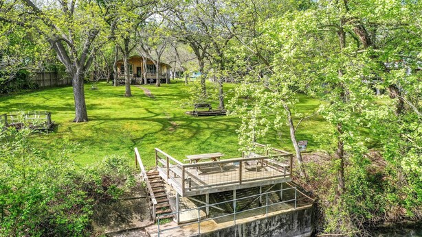 Una terraza de madera con mesas de picnic que da a un río, rodeada de exuberante vegetación y árboles.