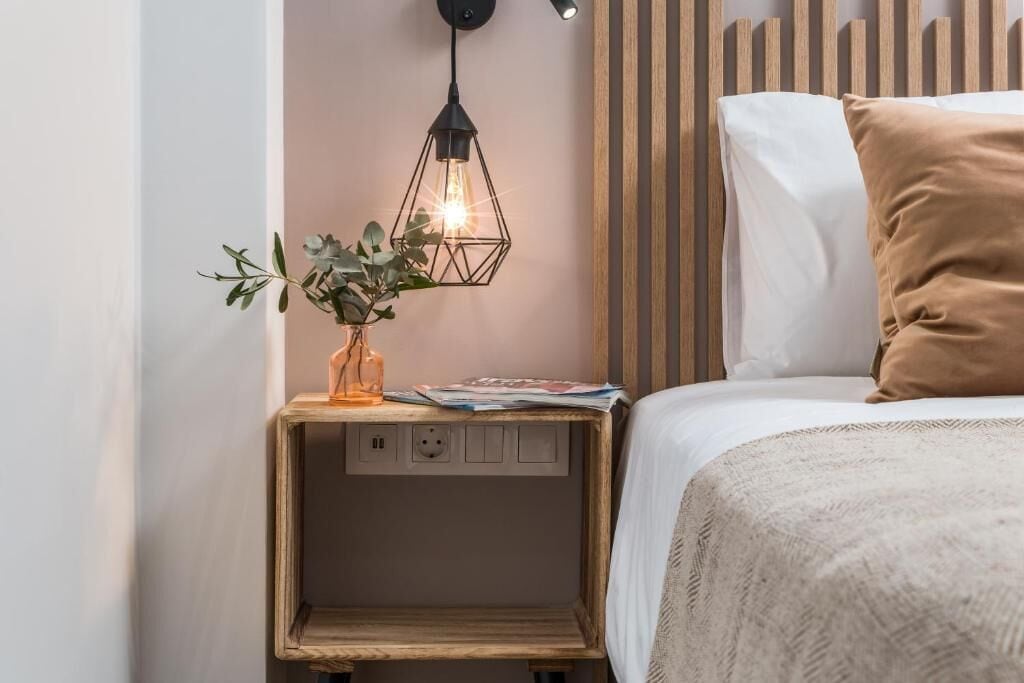 Una cama bien hecha con un almohadón blanco y marrón, una mesita de noche con una planta y una lámpara, y un aplique de pared.