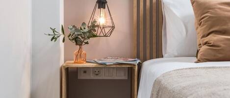 Una cama bien hecha con un almohadón blanco y marrón, una mesita de noche con una planta y una lámpara, y un aplique de pared.