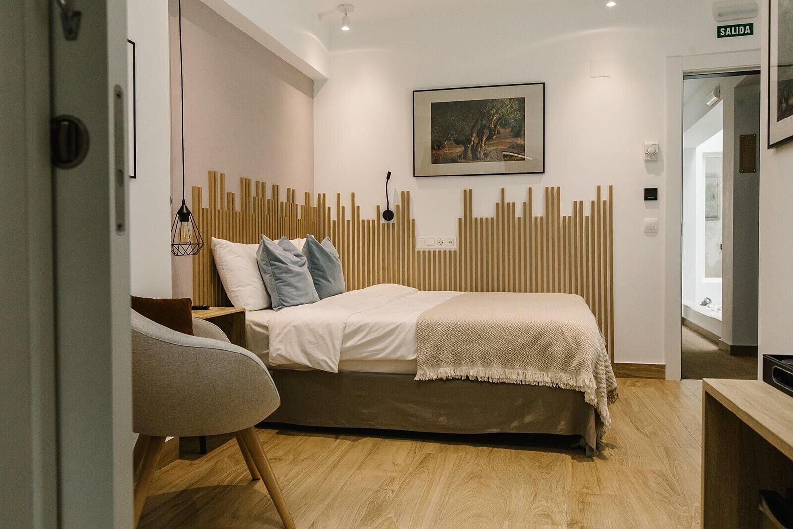 A modern bedroom with a wooden headboard, a bed with white linens, a gray chair, and a framed picture on the wall.