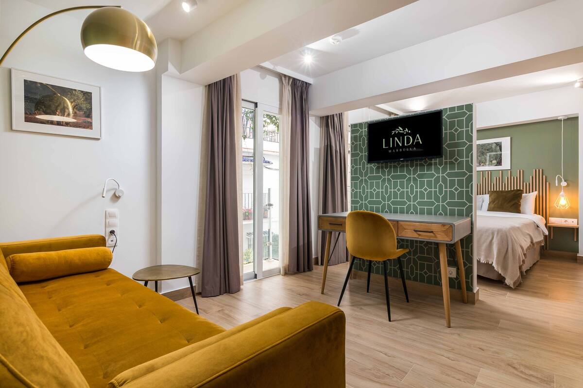 A modern hotel room with a sofa, a desk, a TV, and a balcony with curtains.