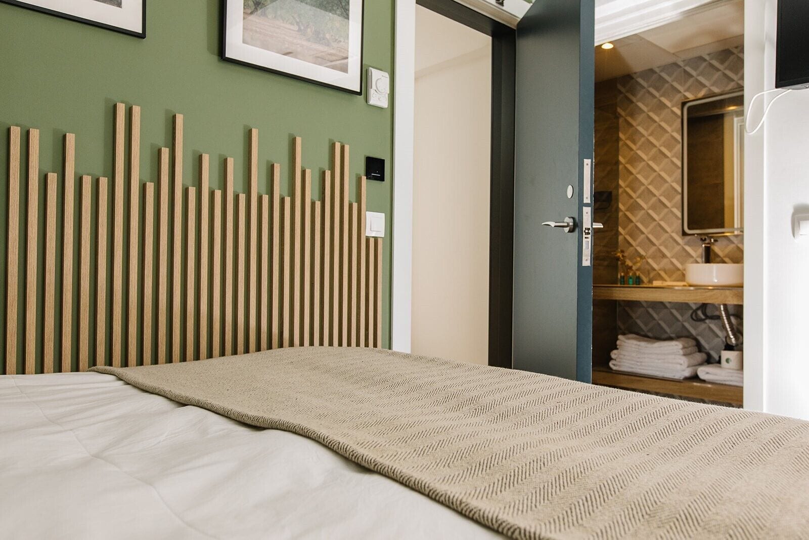 A bedroom with a bed, a wooden slat headboard, a bathroom with a sink and mirror, and a towel rack.