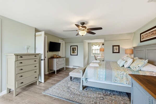 A bedroom with a bed, dresser, ceiling fan, and a TV on the wall.