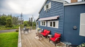 A blue building with a wooden deck, outdoor seating, and a view of the surrounding greenery. - Cozy cottage with a hot tub on the bay of fundy (Berwick)