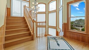 Una escalera de madera con un candelabro, ventanas amplias con persianas y vista a una casa.