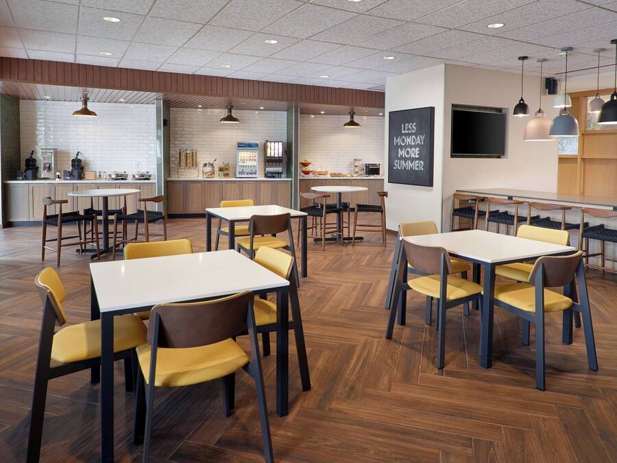 A modern dining area with multiple tables and chairs, a kitchen in the background, and a motivational poster on the wall.