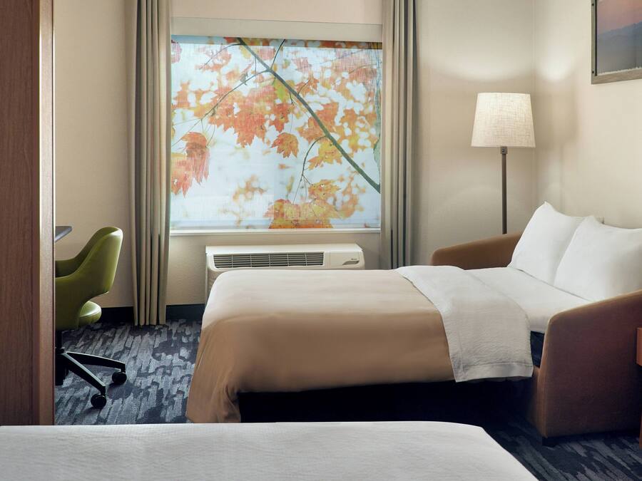 A hotel room with a bed, a chair, a desk, a lamp, and a window with a view of autumn leaves.