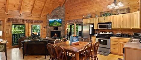 A wooden cabin interior with a dining area, kitchen, fireplace, and a TV mounted on the wall.
