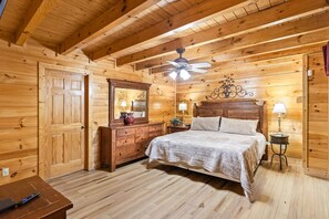 A wooden cabin bedroom with a bed, dresser, mirror, nightstands, lamps, and a ceiling fan.