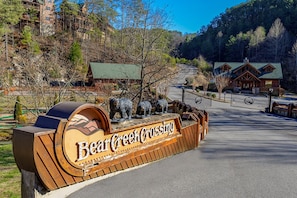 Señal de madera de Bear Creek Crossing con estatuas de oso, una carretera pavimentada y un área boscosa con edificios al fondo.