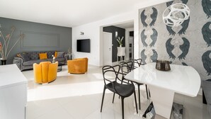 Een moderne woonkamer met een grijze bank, een geel poefje en een witte eettafel met zwarte stoelen.