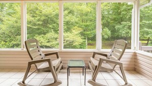 Ein Zimmer mit zwei Schaukelstühlen und einem kleinen Tisch, große Fenster mit Blick auf einen Wald.