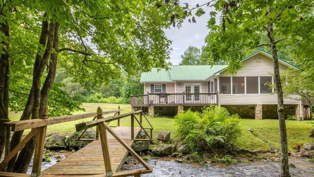 Eine Holzbrücke über einen Bach, der zu einem Haus mit grüner Dachdeckung und Steinmauerwerk führt.
