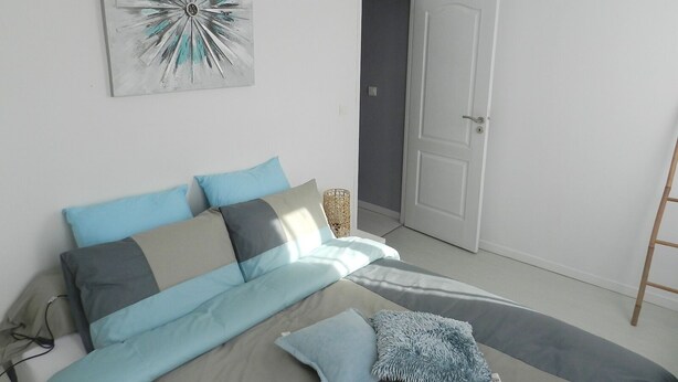 A bedroom with a bed, pillows, a blanket, a wall art piece, a door, and a hanging light fixture. - The blue house (Fécamp)