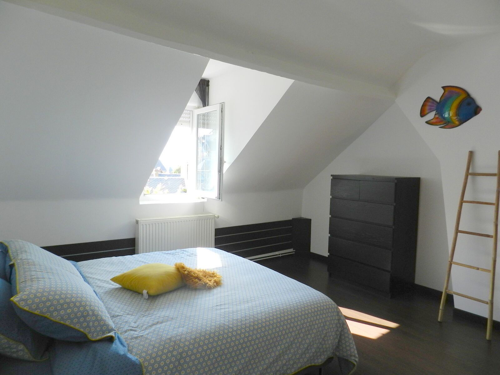 A bedroom with a sloped ceiling, a bed with patterned bedding, a radiator, a dark dresser, and a ladder against the wall.
