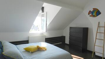 A bedroom with a sloped ceiling, a bed with patterned bedding, a radiator, a dark dresser, and a ladder against the wall.
