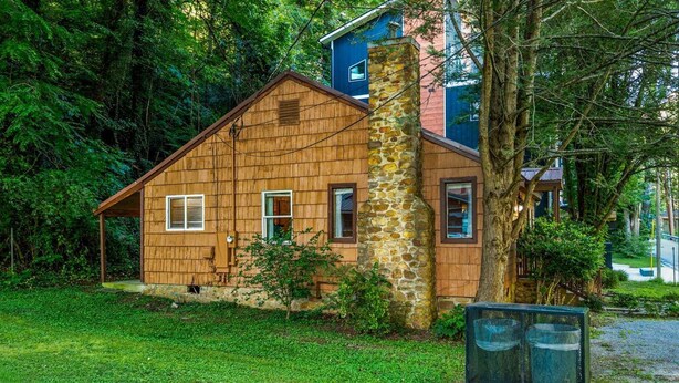 A wooden cabin with a stone chimney, surrounded by lush greenery and a small fenced area.