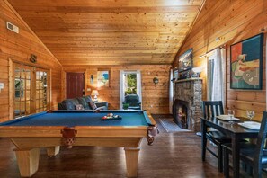 Interior de una cabaña de madera con mesa de billar, chimenea y comedor.