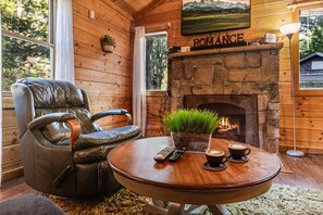Ein gemütliches Zimmer mit Kamin aus Stein, Ledersessel und rundem Holztisch mit Pflanze und zwei Tassen Kaffee.