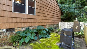 A wooden house with a stone foundation, a window, and a barbecue grill in the yard.