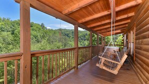 Eine Holzterrasse mit einem Schaukelstuhl, umgeben von einem Hollerleger, mit Blick auf ein bewaldetes Gebiet.