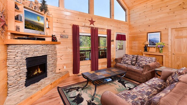 Ein gemütliches Blockhaus-Interieur mit Kamin aus Stein, einer Couch, einem Fernseher und einem Fenster mit Blick auf Grünflächen.