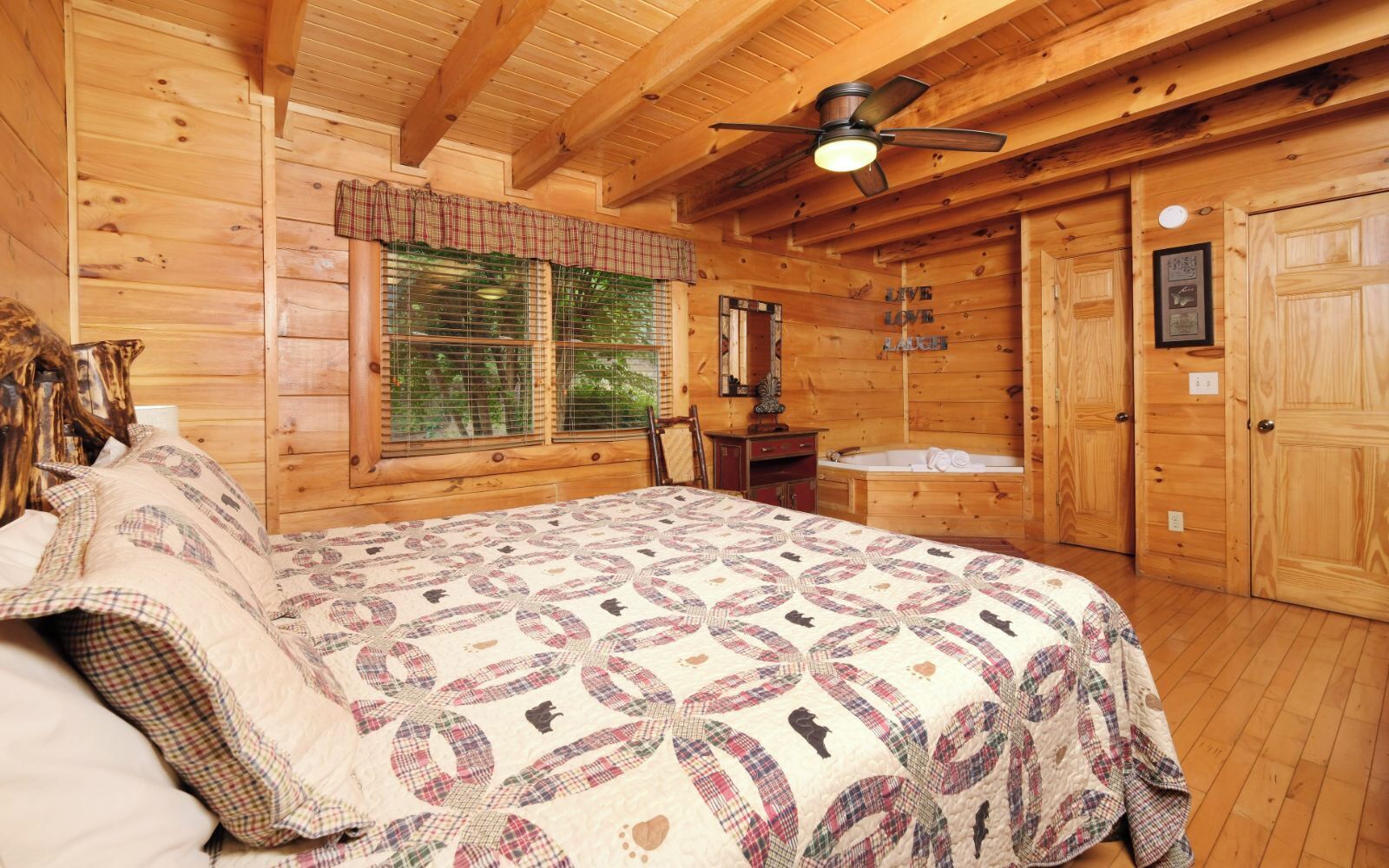A wooden cabin bedroom with a large bed, a ceiling fan, a window with blinds, a small wooden cabinet, and a door.