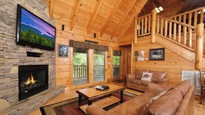 A cozy cabin interior with a stone fireplace, a wooden staircase, and a leather sofa.