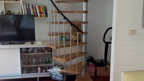A living room with a wooden staircase, a flat-screen TV, a bookshelf, and a patterned armchair.