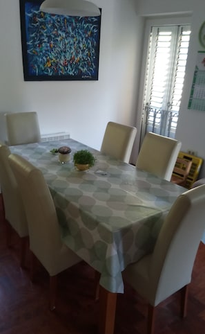 Uma mesa de jantar com toalha de mesa estampada, rodeada por cadeiras. Há uma planta em vaso e um objeto decorativo sobre a mesa.