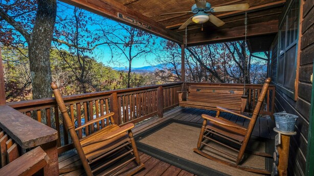 Eine hölzerne Veranda mit Schaukelstühlen und einer Bank, mit Blick auf eine bewaldete Gegend und einen entfernten Berg.