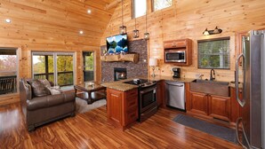 A cozy cabin-style kitchen with wooden floors, a fireplace, a large TV, and a refrigerator.