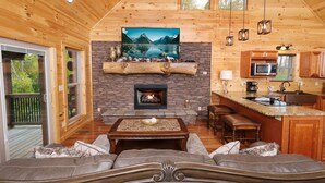 A cozy cabin interior with a fireplace, wooden walls, a stone fireplace surround, a dining area, and a kitchen with modern appliances.