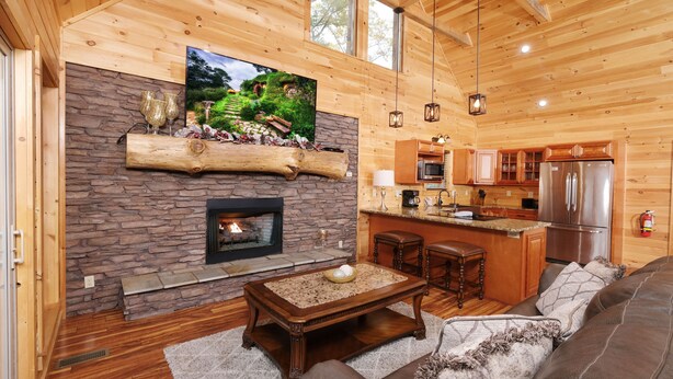 A cozy cabin interior with a stone fireplace, wooden walls, a TV mounted above it, a kitchen area, and a comfortable seating area with a sofa and a coffee table.