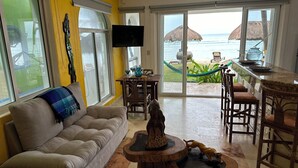 A living room with a sofa, a coffee table, and a TV. There is a view of the ocean through large windows. - Beautiful pool side ground floor unit with direct beach access , WiFi and AC. (Akumal)