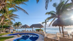A pool area with a clear view of the ocean, surrounded by palm trees and thatched umbrellas. - Beautiful pool side ground floor unit with direct beach access , WiFi and AC. (Akumal)
