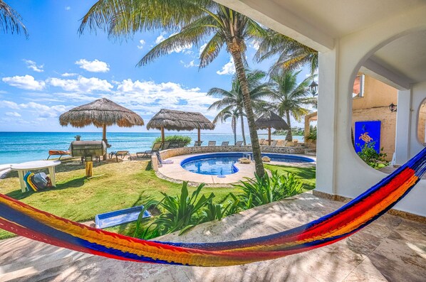 A pool area with a hammock, lounge chairs, and thatched umbrellas overlooking the ocean. - Beautiful pool side ground floor unit with direct beach access , WiFi and AC. (Akumal)