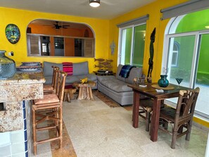 A bright yellow living area with a sofa, wooden chairs, a dining table, and a kitchen area with a counter and stools. - Beautiful pool side ground floor unit with direct beach access , WiFi and AC. (Akumal)