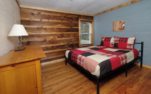 A bedroom with a wooden wall, a bed with a red and black quilt, a wooden nightstand, and a window with blinds.