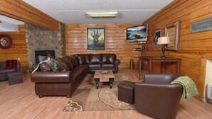 A cozy log cabin interior with a leather sectional sofa, a fireplace, a television, and a family sign on the wall.
