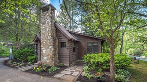A rustic cabin with a stone chimney, surrounded by lush greenery and a small garden.