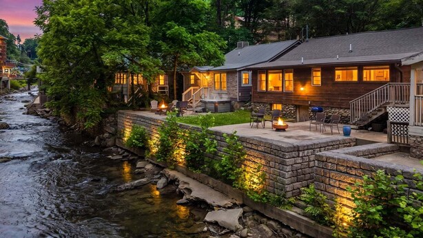 Una cabaña junto al río con un muro de contención de piedra, mobiliario exterior y una fogata.