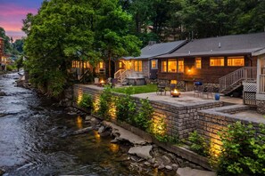Una cabaña junto al río con un muro de contención de piedra, mobiliario exterior y una fogata.