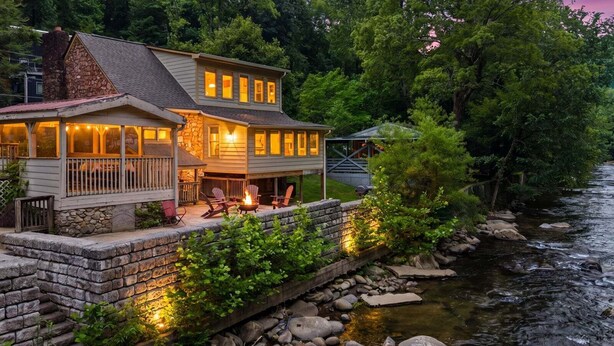 Una casa de dos pisos con base de piedra, un balcón con mobiliario exterior y un río al fondo.
