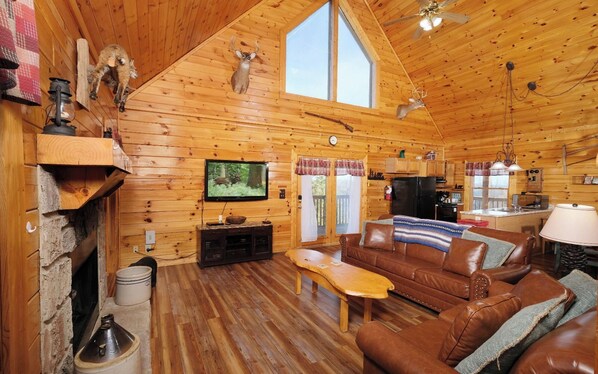 A cozy cabin interior with a wooden interior, a fireplace, a television, a leather sofa, and a kitchen area. - Pioneer Place - Arcade Games, Mountain Views, Hot Tub (Sevierville)