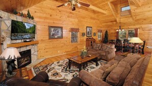 Interior acogedor de una cabaña de troncos con chimenea de piedra, muebles de cuero y un televisor montado en la pared.