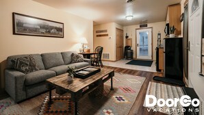 Uma sala de estar com sofá, mesa de centro e um quadro na parede. Há uma porta que dá para a cozinha.